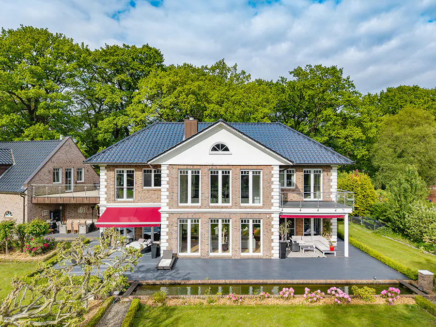 Two-story brick house with a blue roof, white trim, and a red awning, surrounded by green trees and a lawn.