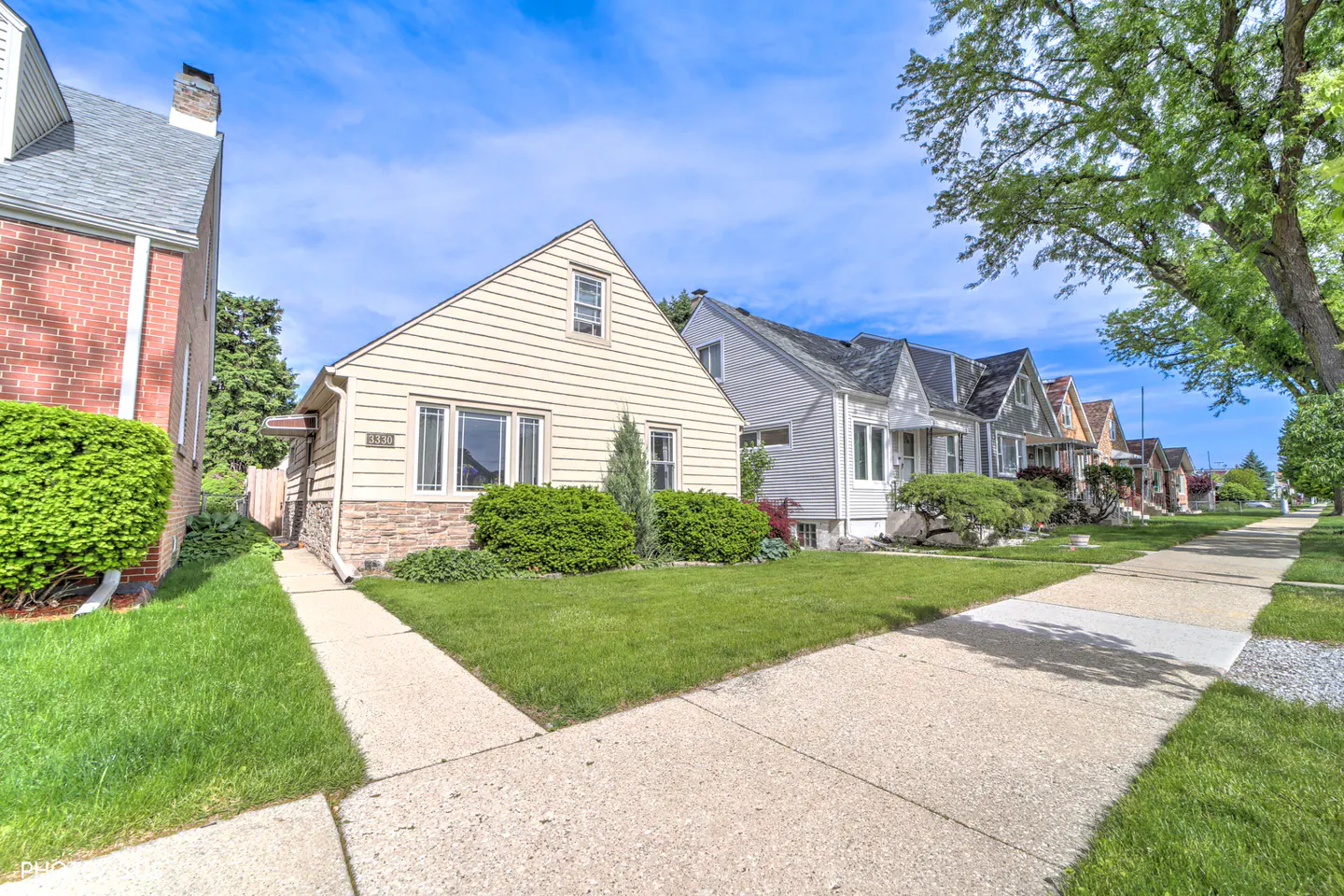 A beige, single-family home with a green lawn and sidewalk on a sunny day. Other houses line the street.