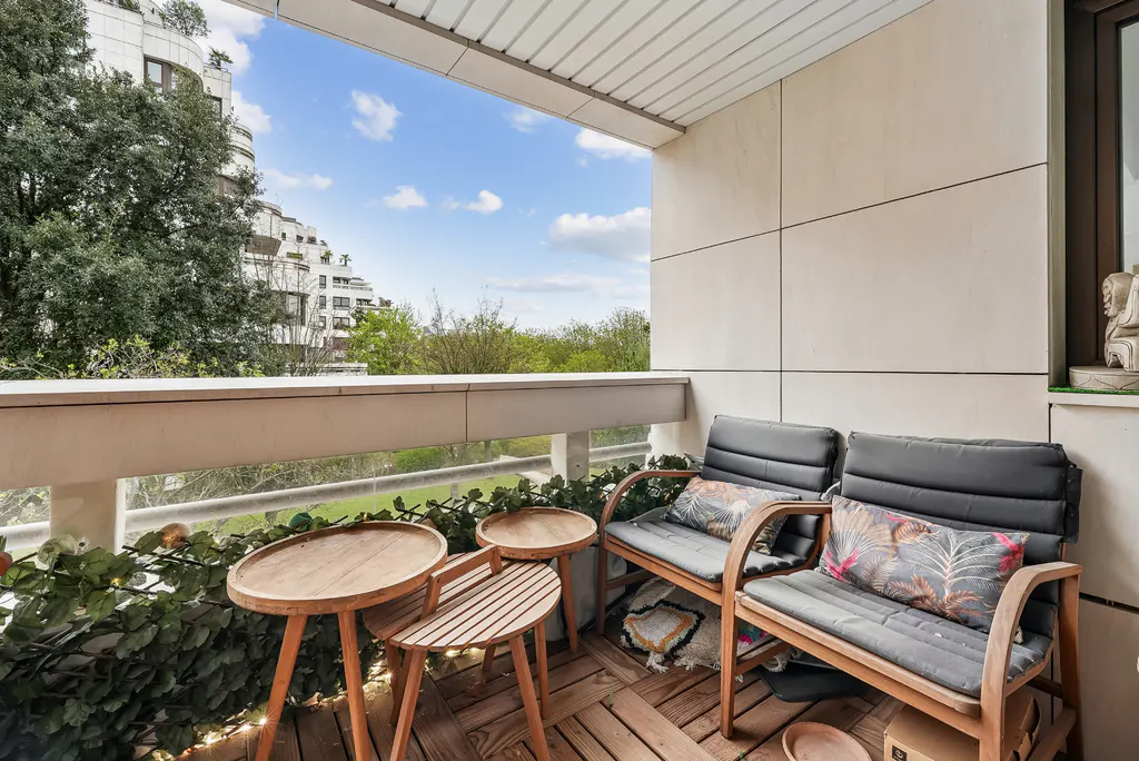 Balcony with two chairs, pillows, and tables on a wooden floor. Outside, trees and a building are visible under a blue sky.