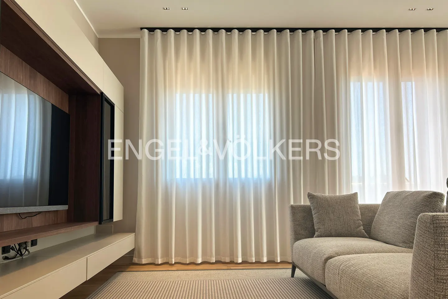 Modern living room with a grey sofa, wall-mounted TV unit, and floor-to-ceiling white curtains covering a large window.