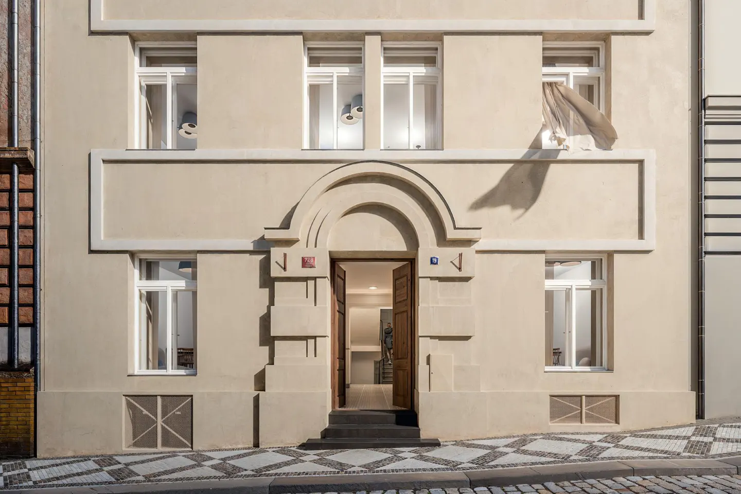 Facade of a beige building with white windows and an open wooden door. A curtain billows from an upper window. Cobblestone street.