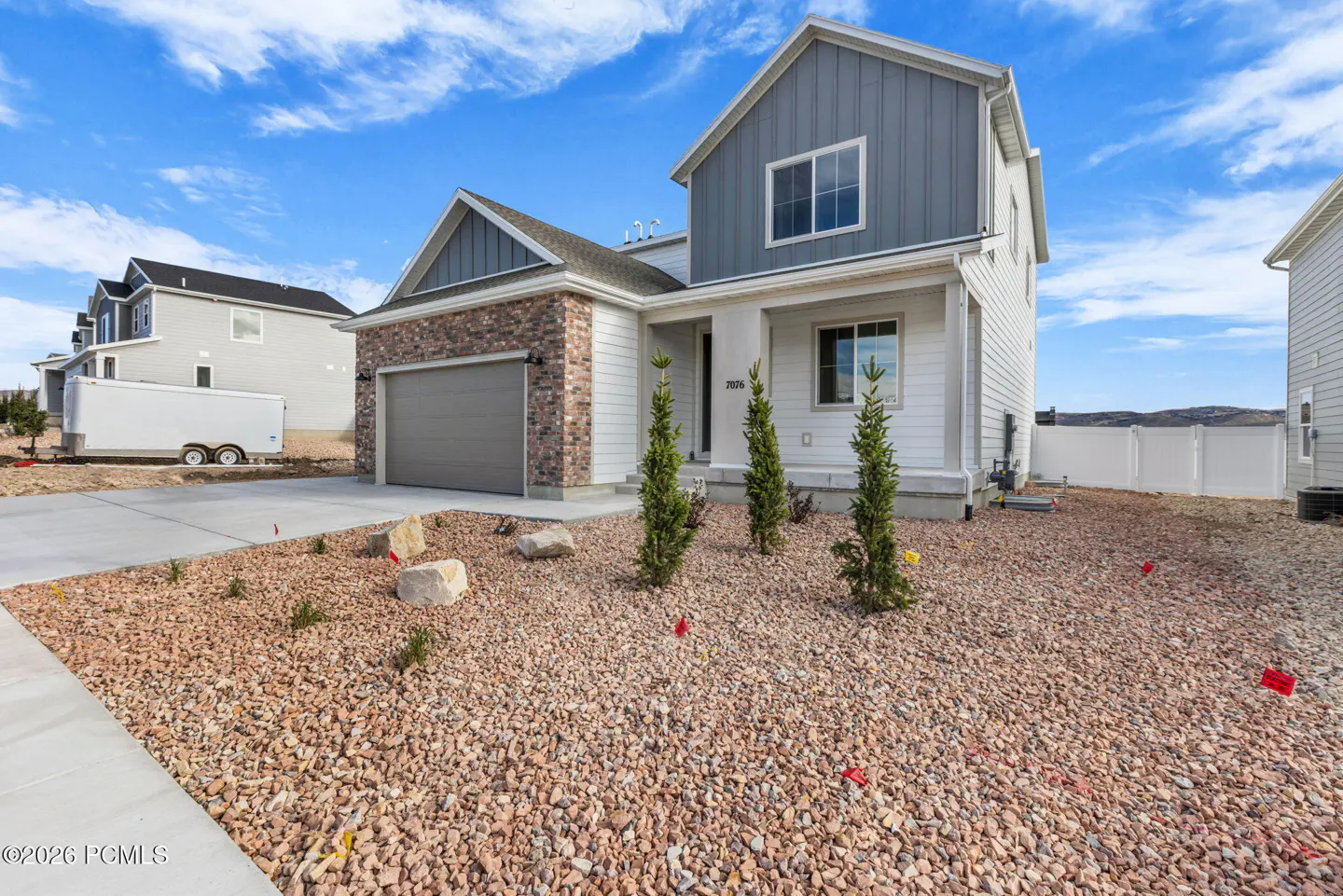 Two-story house with gray and white siding, brick accents, and a gray garage door. The yard is covered in small rocks. Blue sky with clouds.