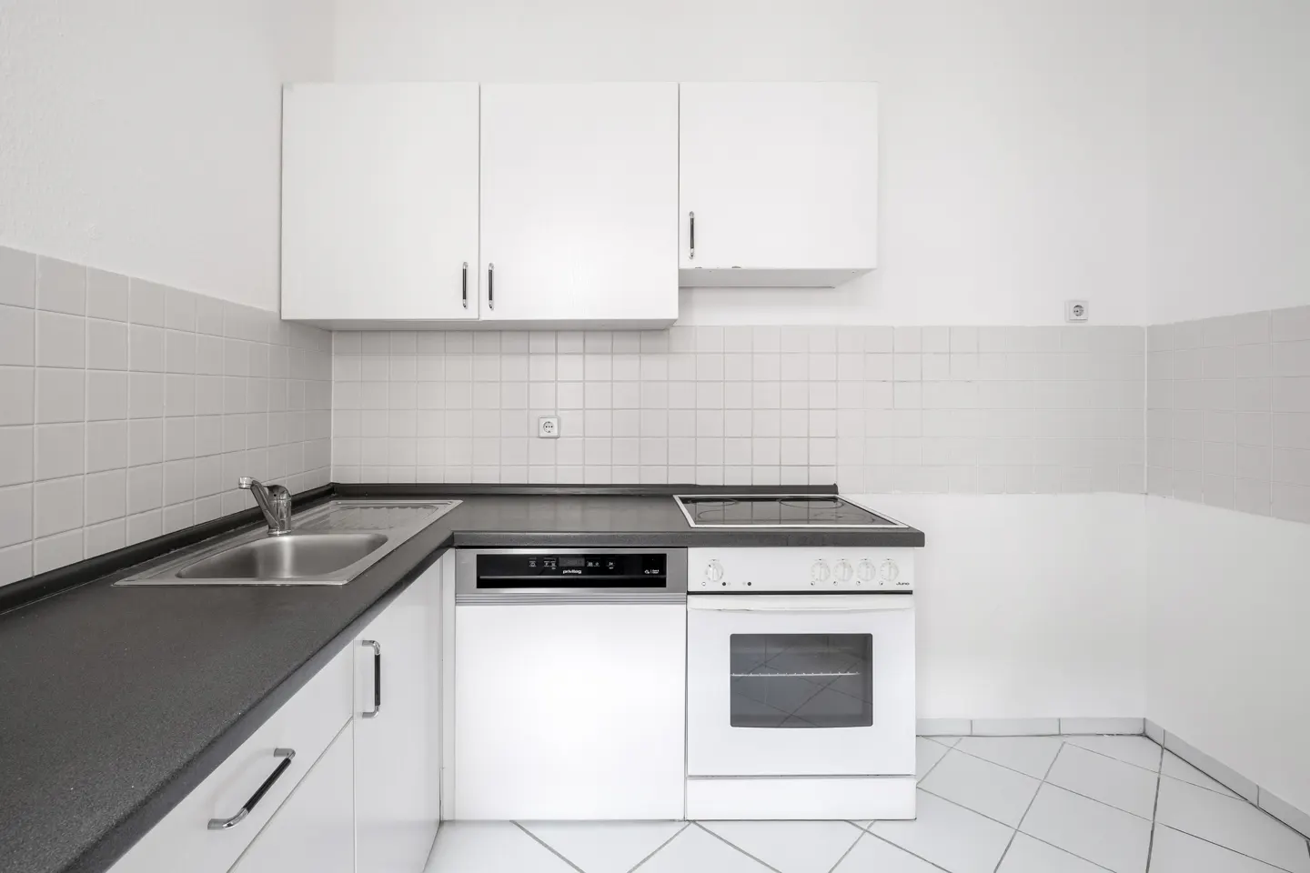 A bright, minimalist kitchen with white cabinets, black countertops, and white tiled walls and floor. Appliances include a sink, dishwasher, and oven.