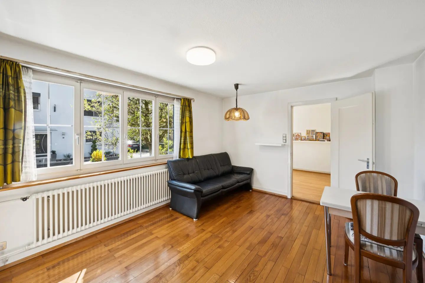 Bright room with wood floors, a black sofa, and a table with two chairs. Large windows with yellow curtains let in natural light.