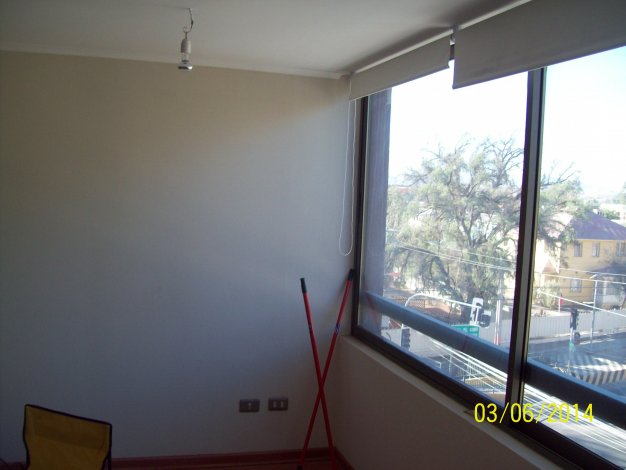 A bright, empty room with a large window showing a street scene with trees and buildings. A yellow chair and red poles are in the corner.