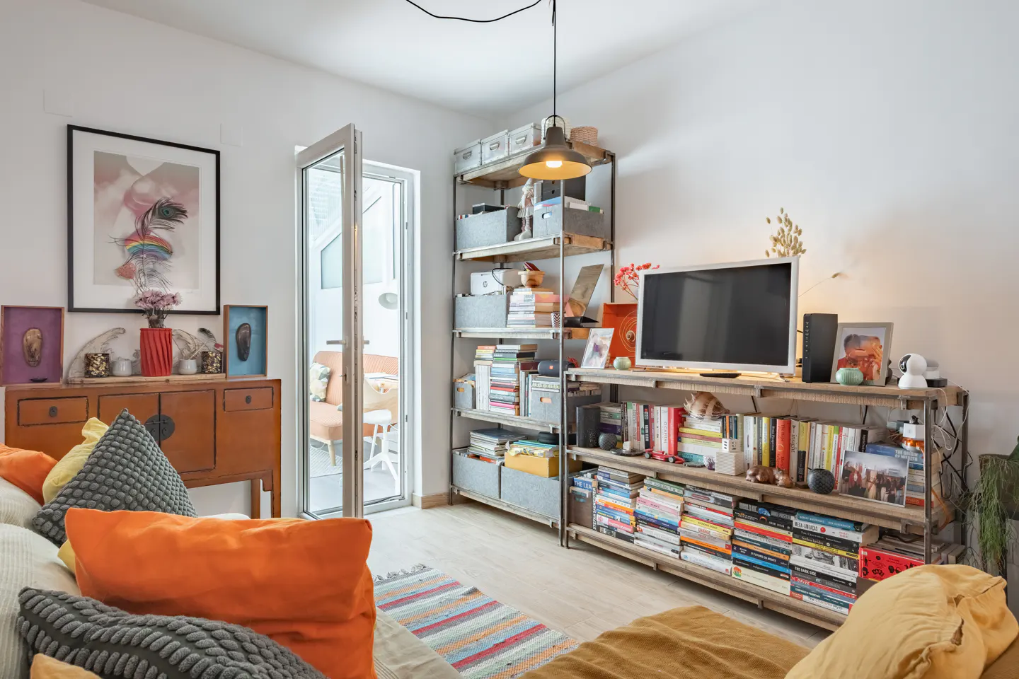 Living room with a brown cabinet, artwork, and shelves filled with books and decor. A glass door leads to an outdoor seating area.