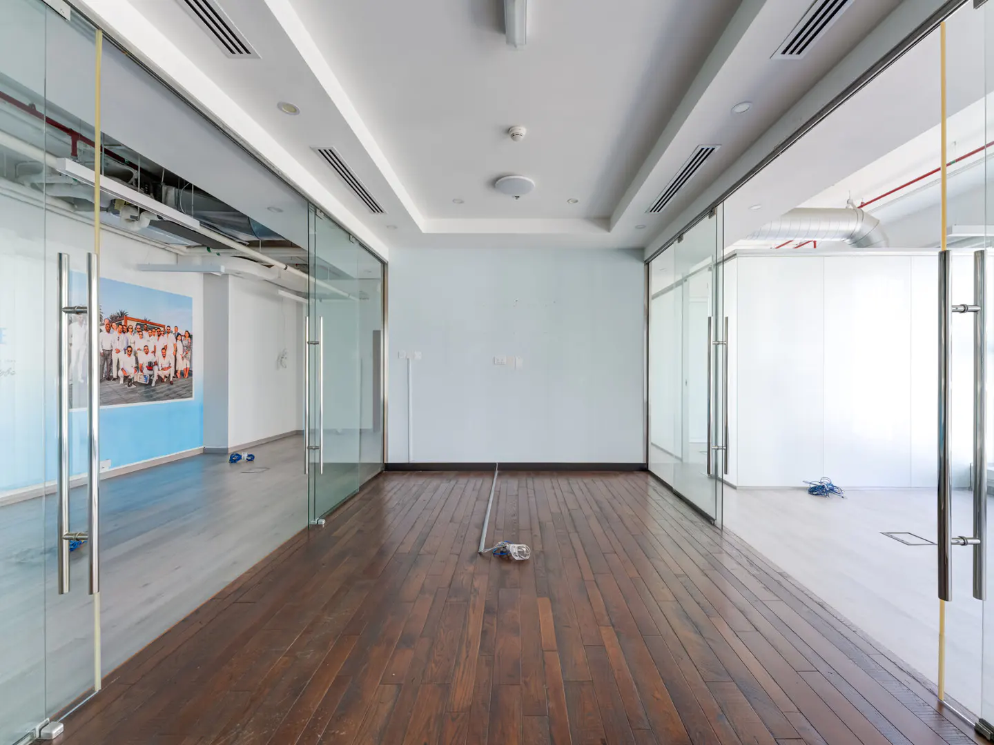 Empty office space with hardwood floors, white walls, and glass-walled rooms on either side.