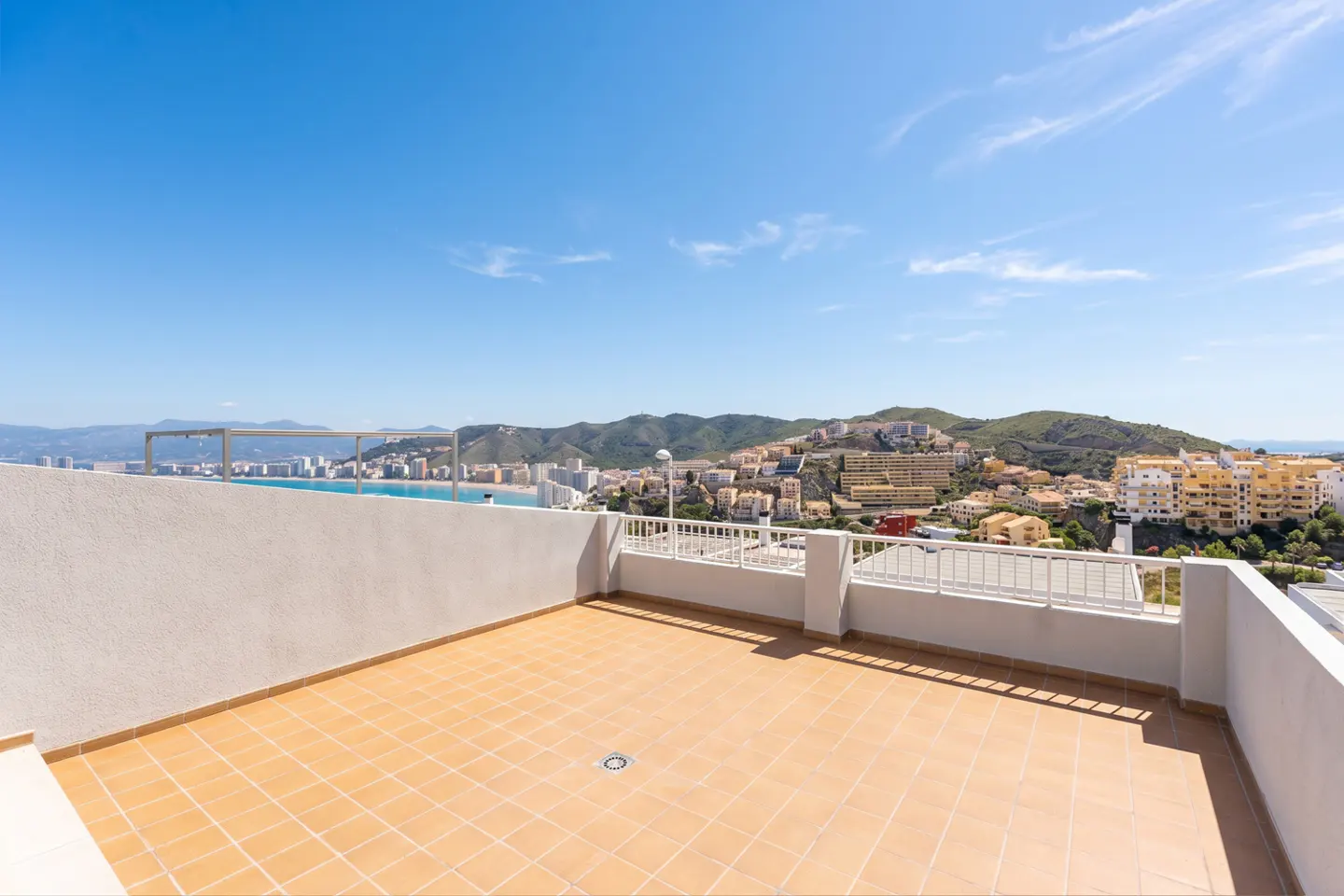 Tiled rooftop terrace with white railings overlooking a coastal city and mountains under a clear blue sky.