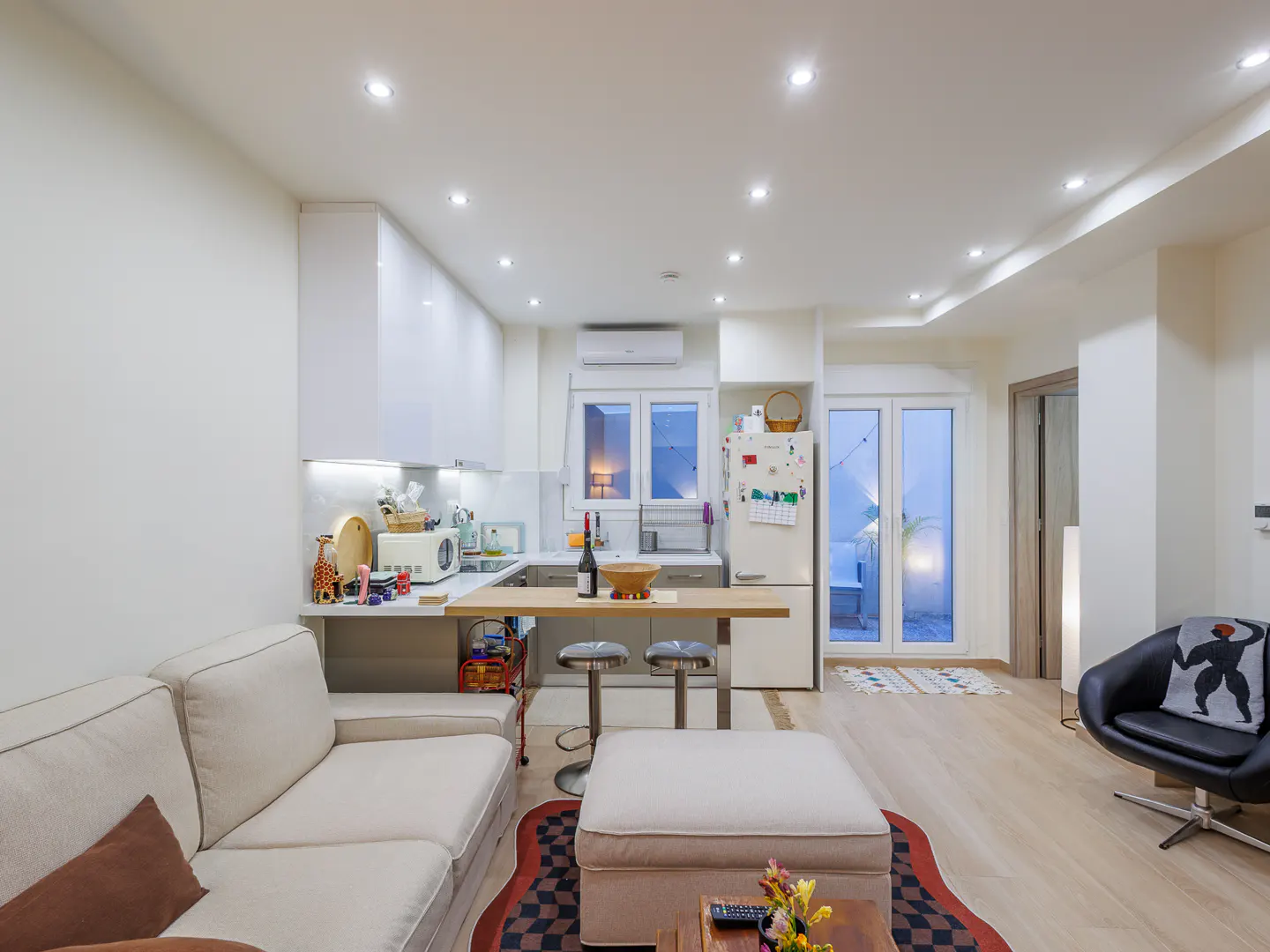 Bright, open-concept living space with white walls, light wood floors, and modern furniture. Kitchen area with white cabinets and island.