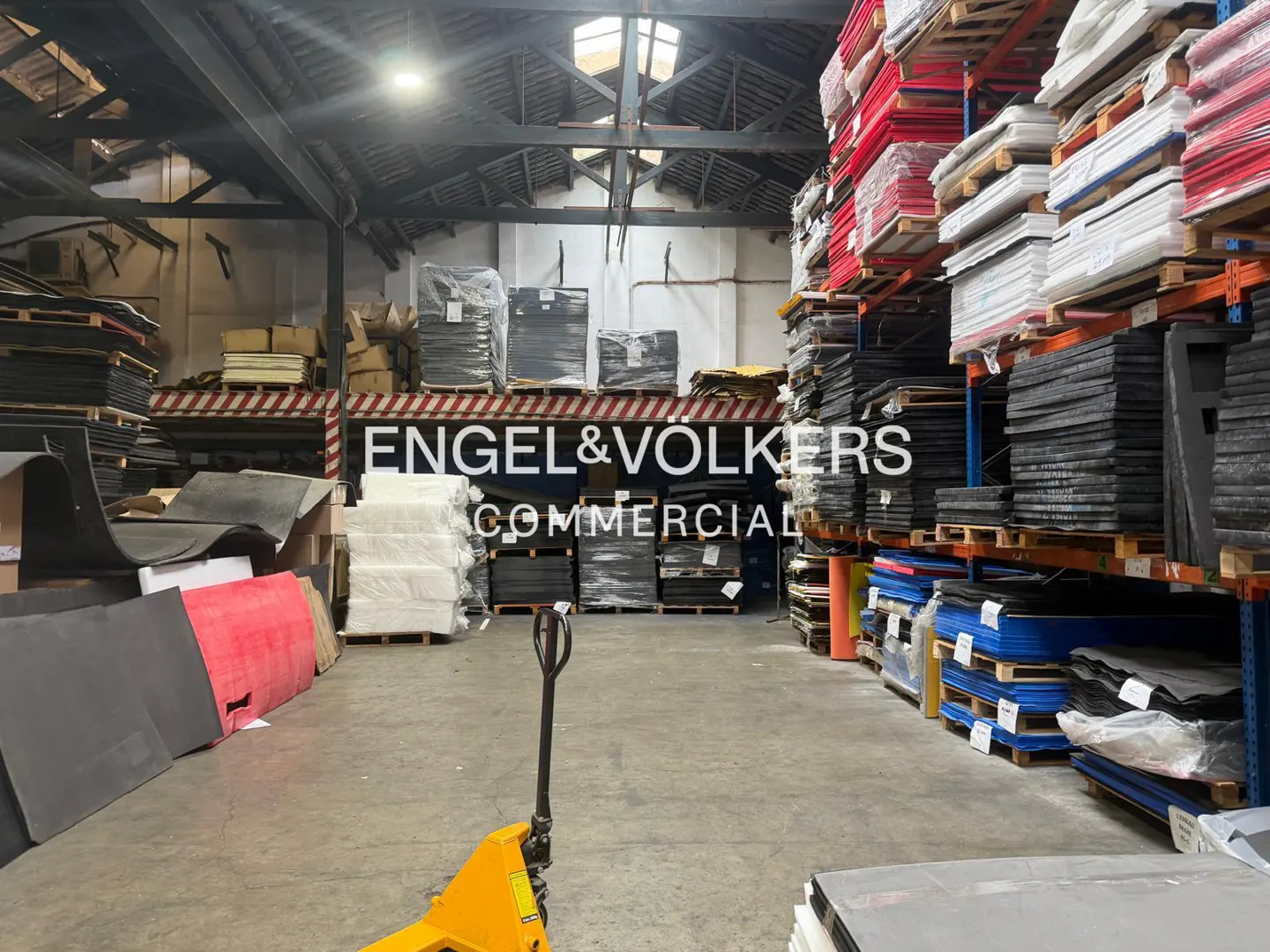 A warehouse interior with stacked materials, a yellow pallet jack, and the Engel & Volkers Commercial logo.