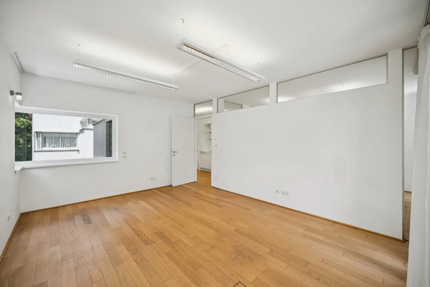 Minimalist white room with light wood floors, fluorescent ceiling lights, and a window looking out to a building exterior.