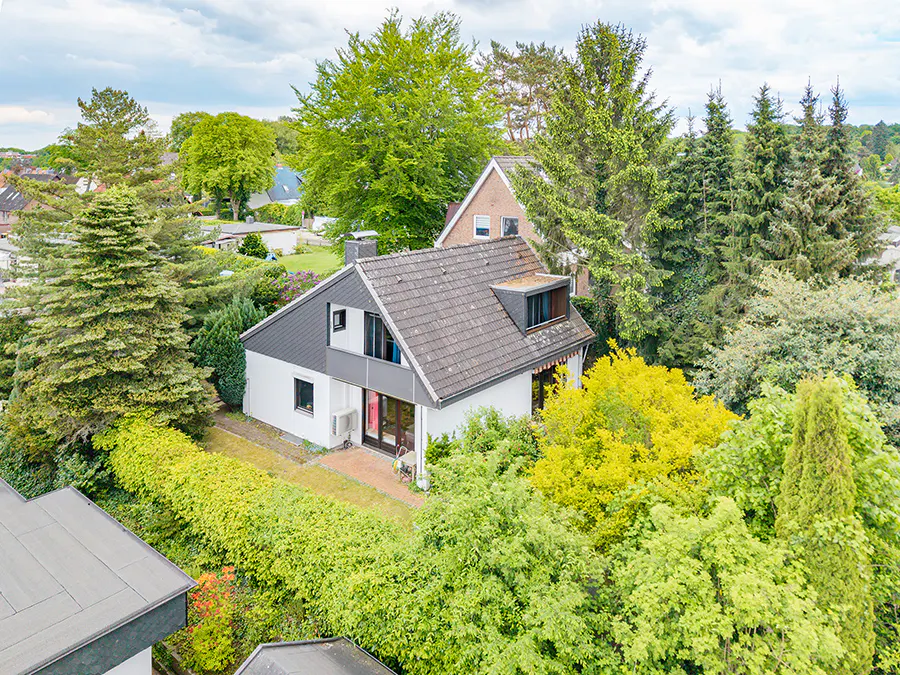 Aerial view of a white house with a gray roof surrounded by lush green trees and bushes.