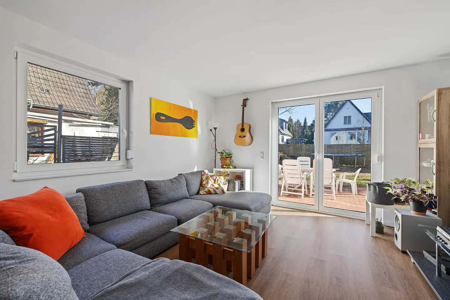 Bright living room with gray sectional, orange pillow, glass coffee table, guitar on wall, and patio doors to backyard.