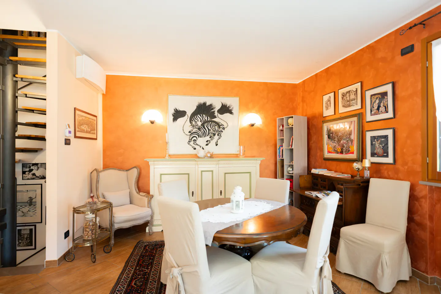 A dining room with orange walls, a wooden table with white chairs, and a zebra painting.
