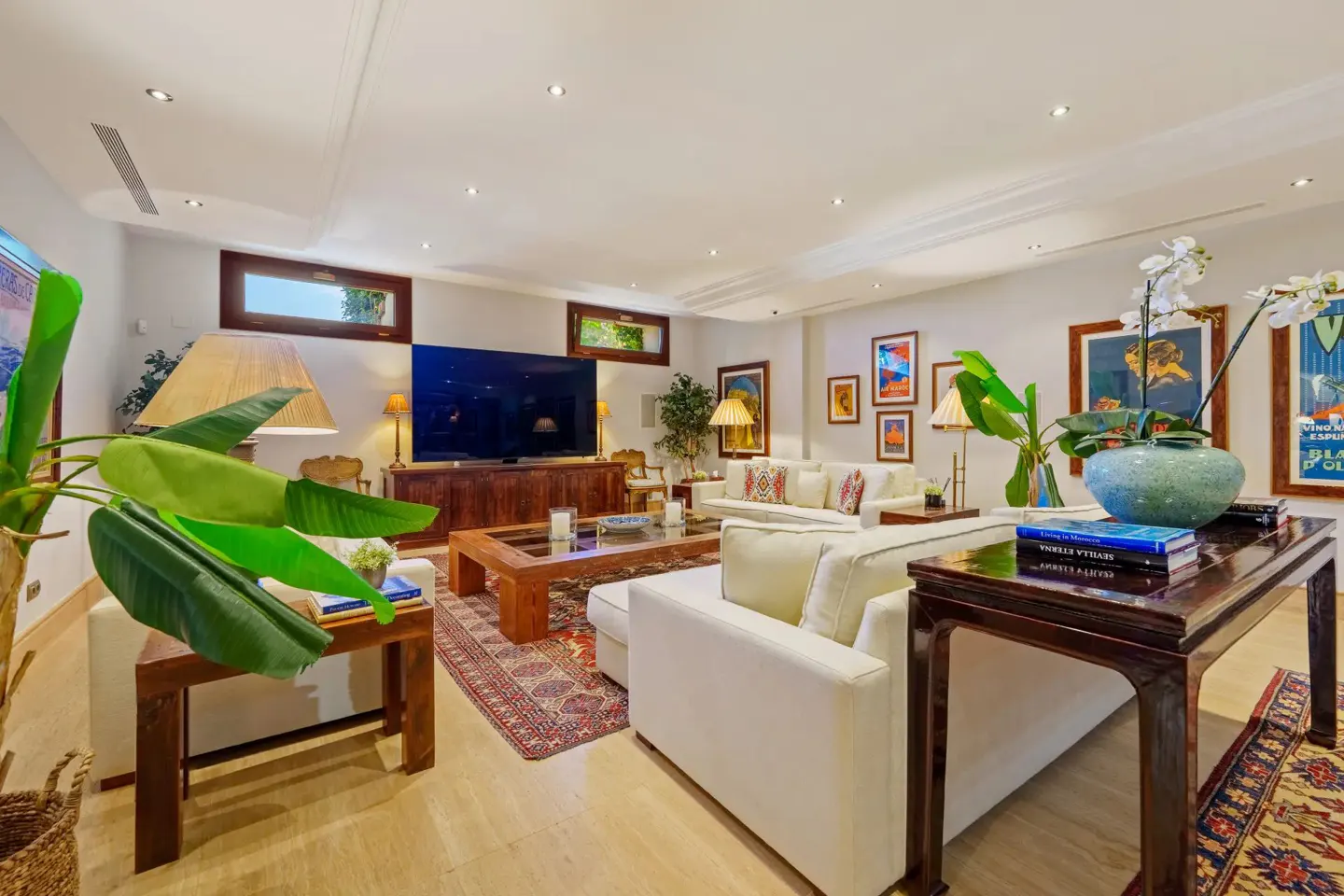 A bright living room with white sofas, wooden tables, and colorful artwork on the walls. A large TV sits above a wooden cabinet.