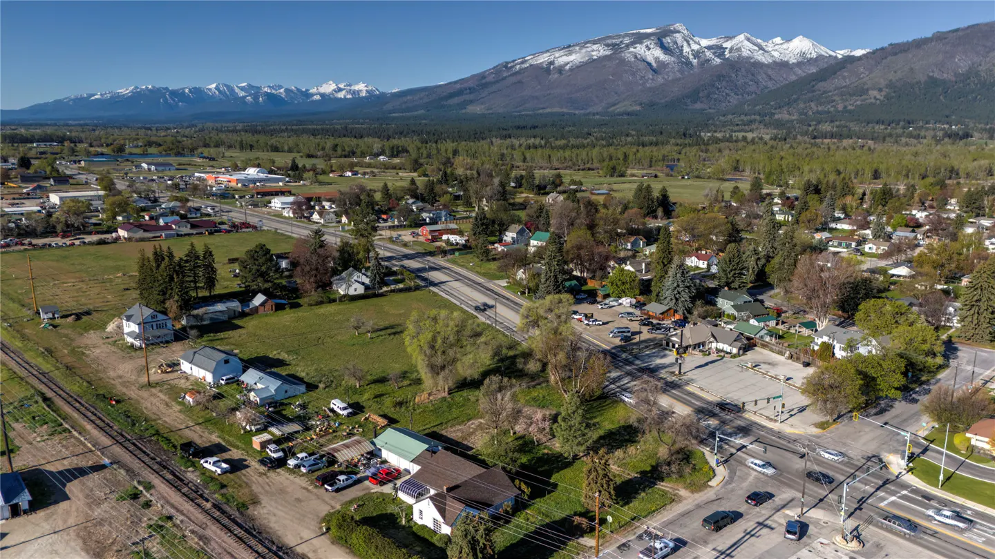 Aerial view of a small town with snow-capped mountains in the background. Houses, trees, and roads are visible.