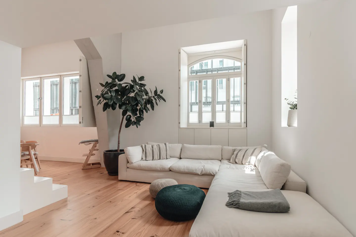 Bright living room with white walls, wood floors, and a large white sectional sofa. A potted ficus tree sits near a window.