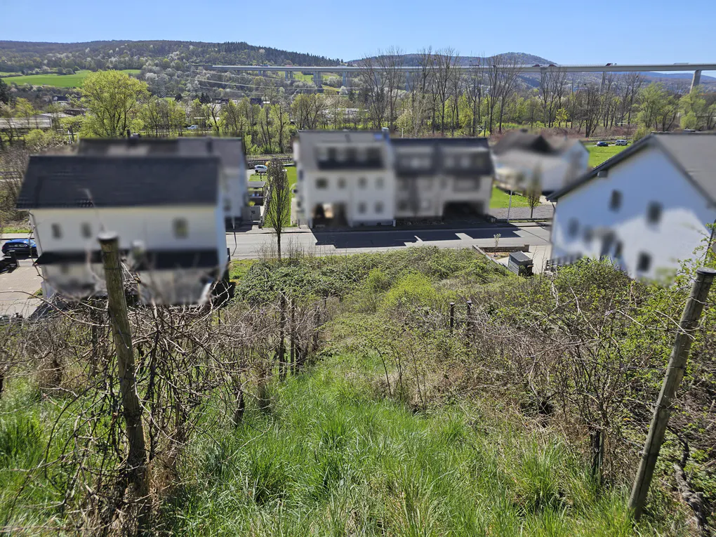 View of a grassy lot with a fence, houses, and a bridge in the background on a sunny day.
