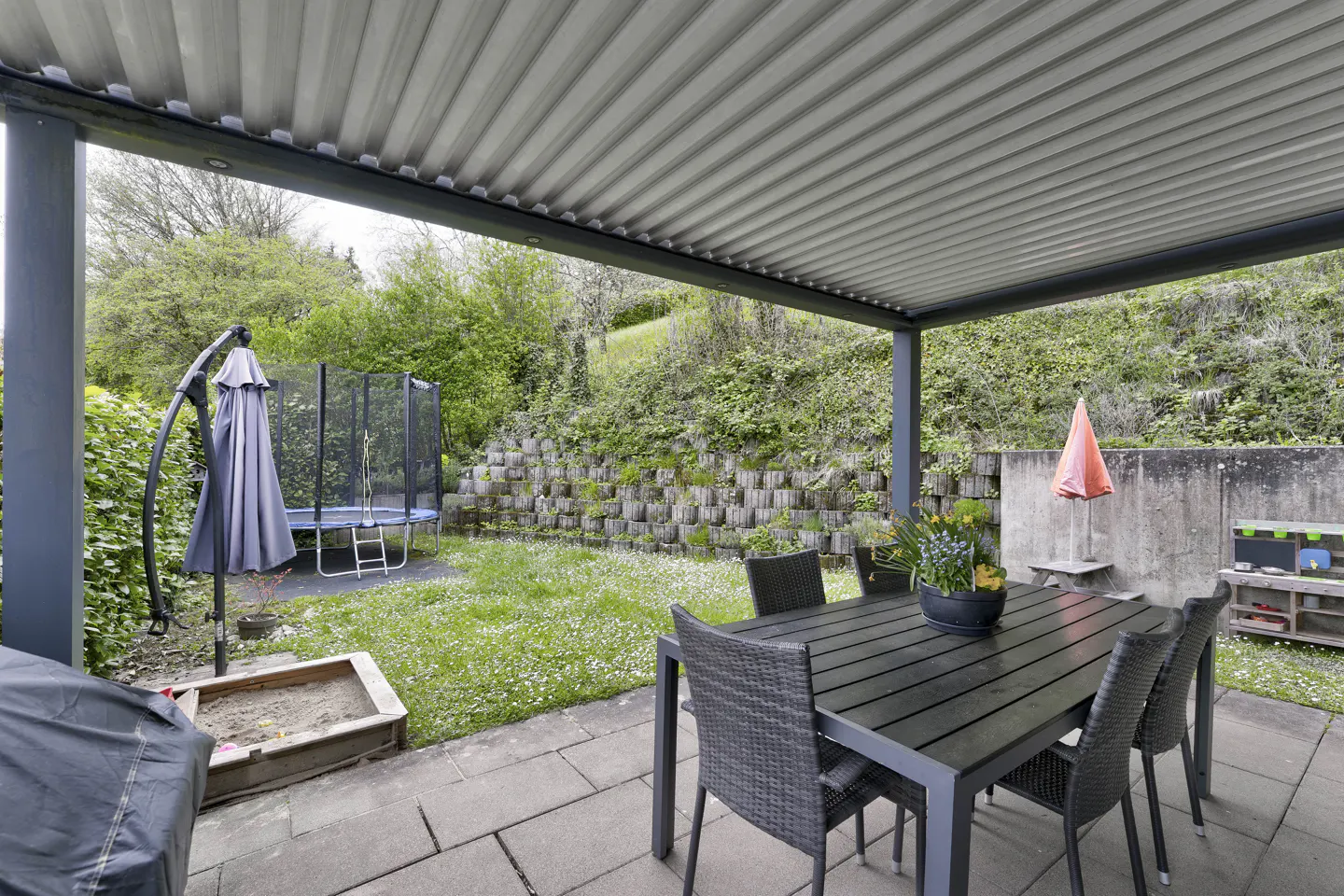 Covered patio with a black table and chairs, a trampoline, and a grassy yard with a retaining wall in the background.