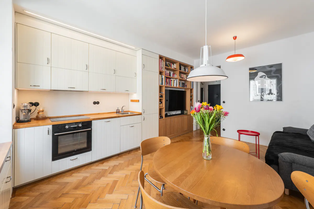 Bright, modern apartment interior with white cabinets, wood countertops, and herringbone floors. A round table with flowers sits in the foreground.