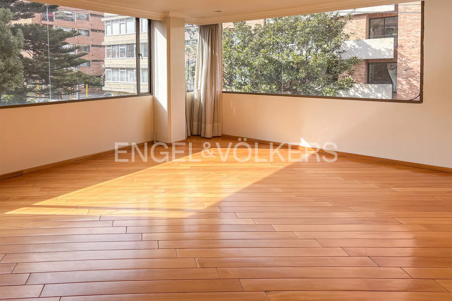 Empty room with light wood floors and white walls. Large windows show trees and buildings. Curtains hang on the right. Sunlight shines on the floor.