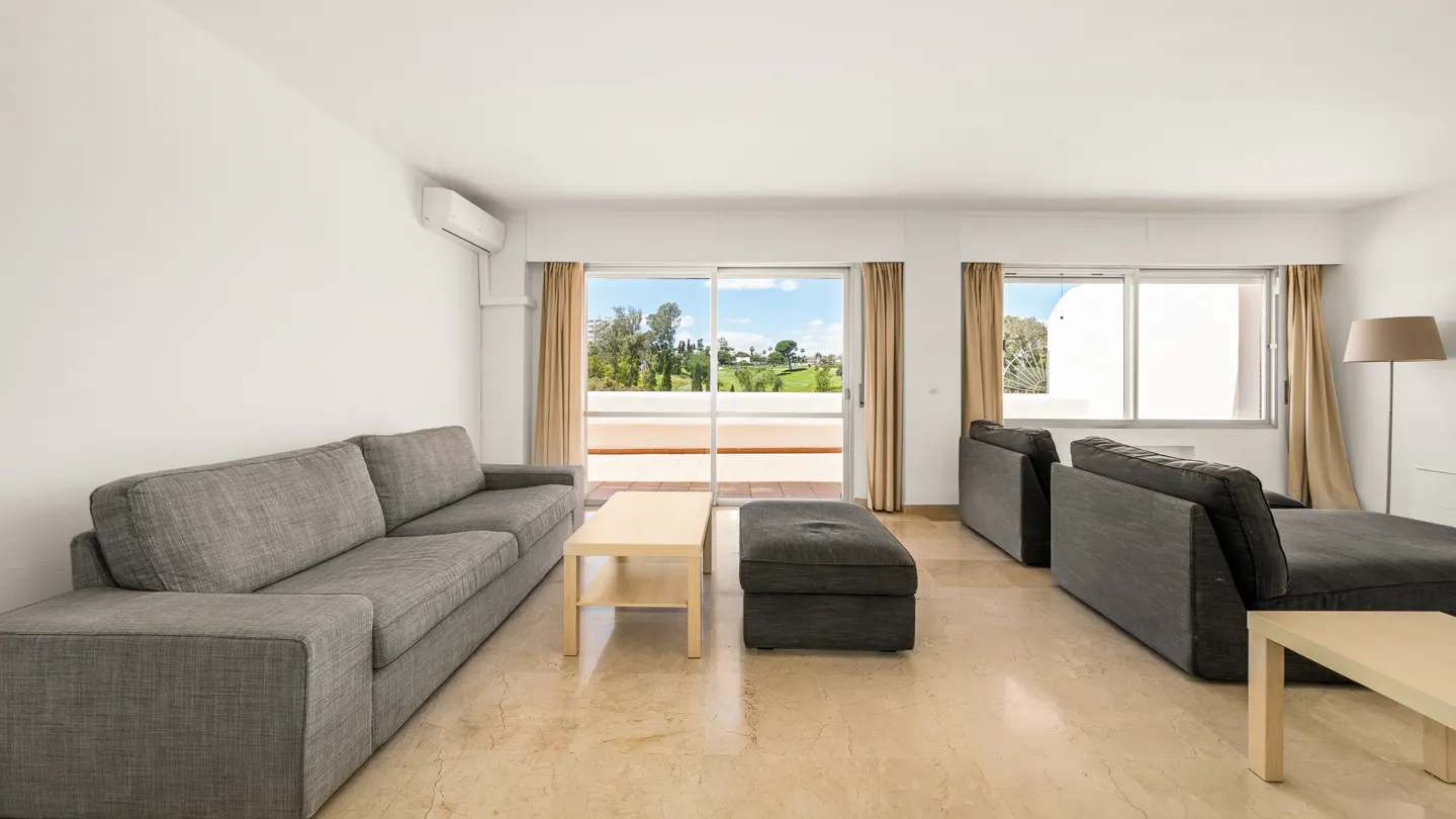 Bright living room with gray sofa, ottoman, and chairs. Sliding glass doors open to a balcony with a golf course view. Beige marble floor.