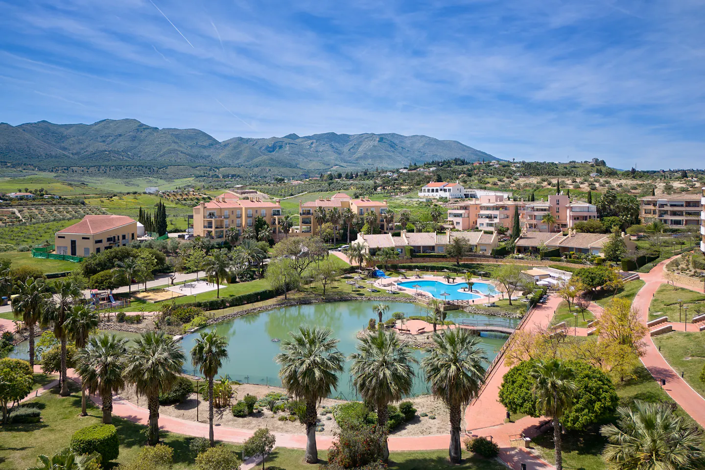 Scenic view of a resort with a lake, pool, and palm trees under a blue sky with mountains in the background.