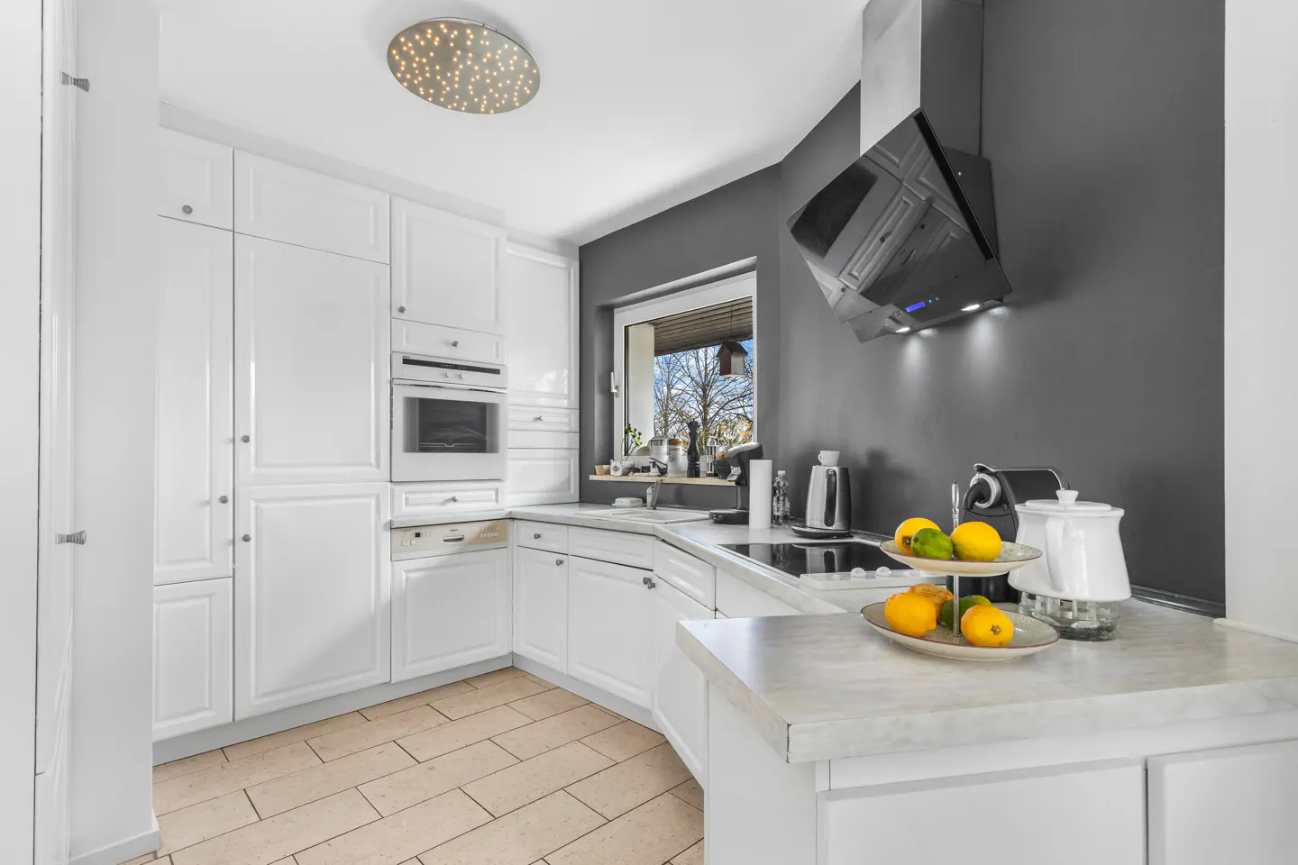 Bright kitchen with white cabinets, beige tile floor, and gray accent wall. A black range hood hangs above the stove. Fruit sits on the counter.