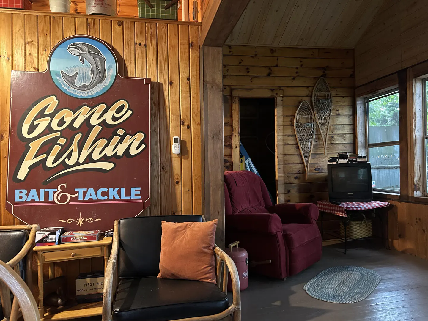 Rustic cabin interior with wood paneling, "Gone Fishin'" sign, red recliner, snowshoes, and vintage TV.