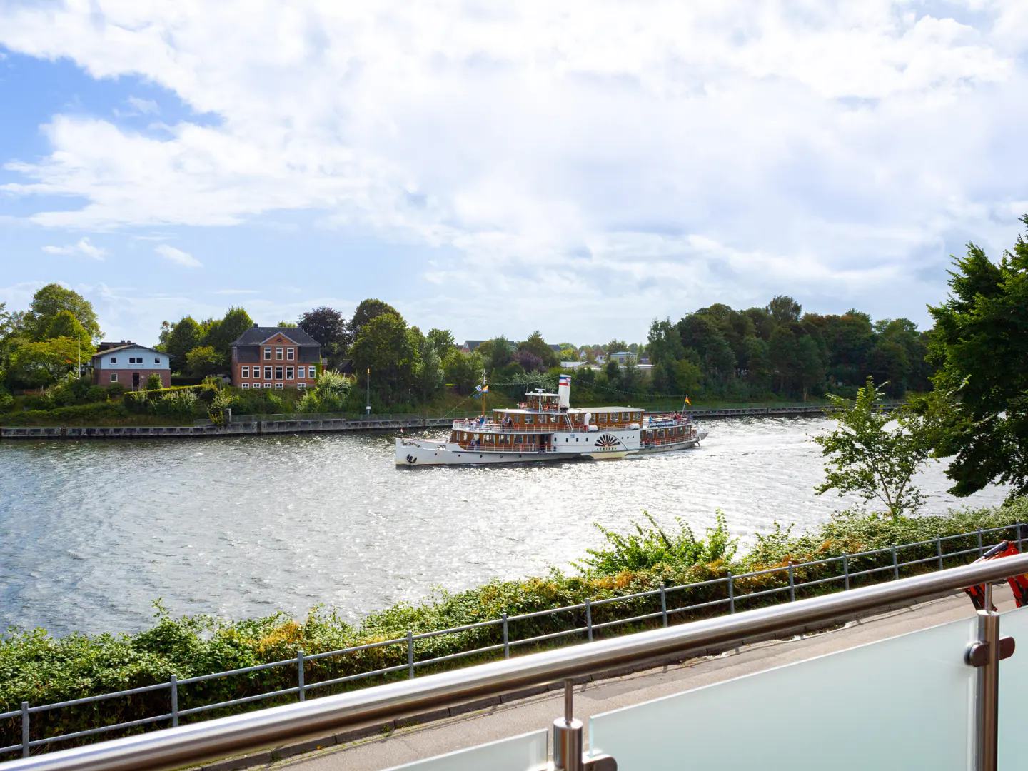 View of a river with a paddle steamer boat, green trees, and houses under a cloudy sky.