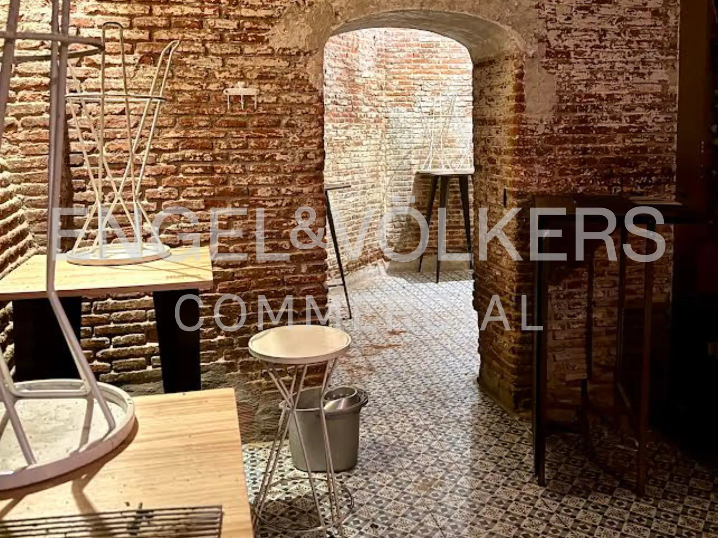 Interior view of a commercial space with exposed brick walls, tiled floors, and arched doorway. Tables and chairs are scattered throughout the room.