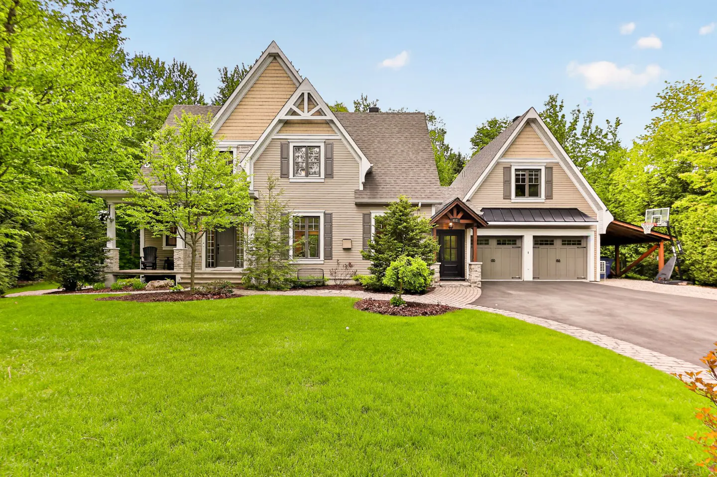 Two-story beige home with a two-car garage, stone accents, and a large green lawn surrounded by lush trees.