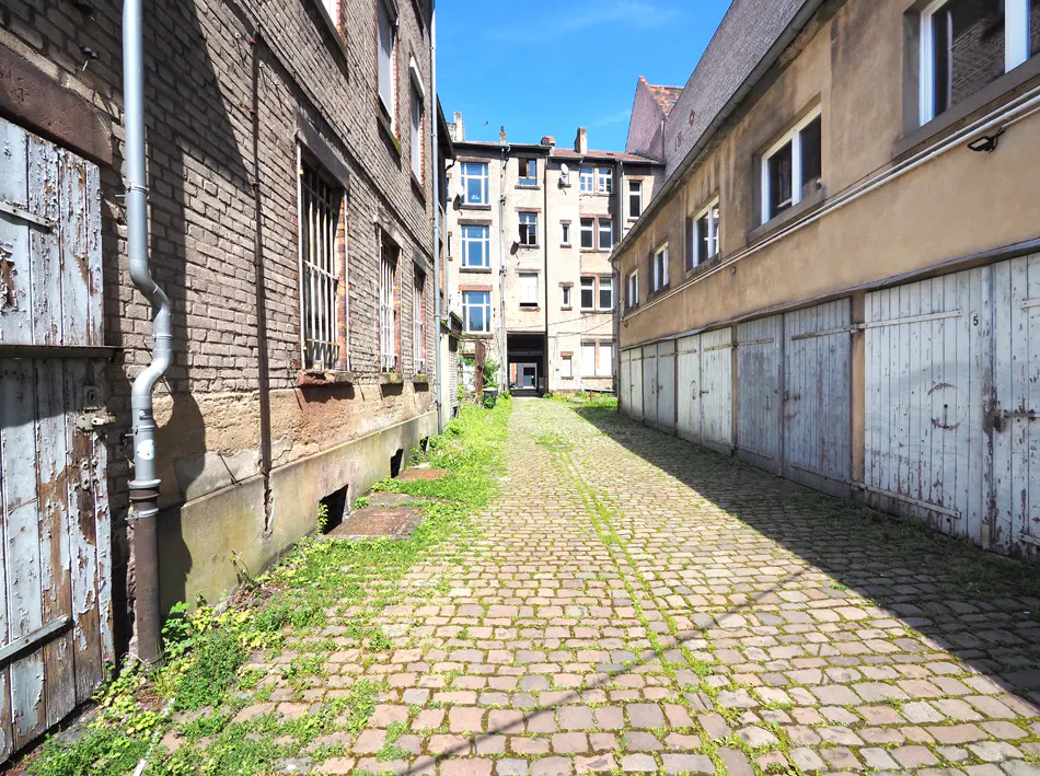 A cobblestone alleyway between brick buildings with peeling paint and weathered garage doors.
