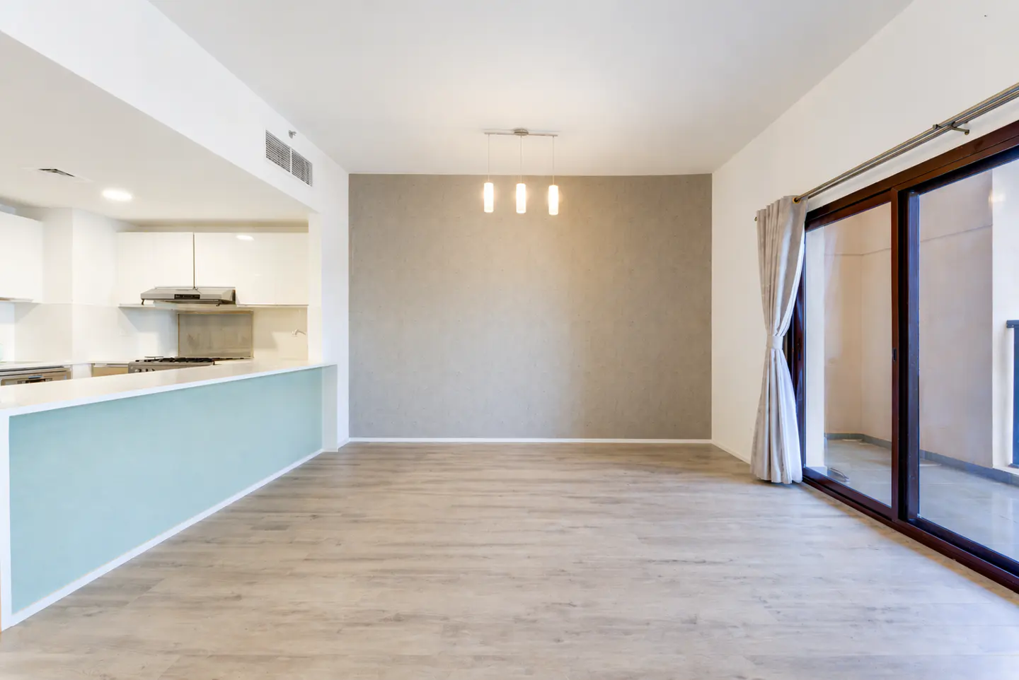 An open-concept living space with light wood floors, a gray accent wall, and a sliding glass door to a balcony. A kitchen with white cabinets and a light blue island is visible.