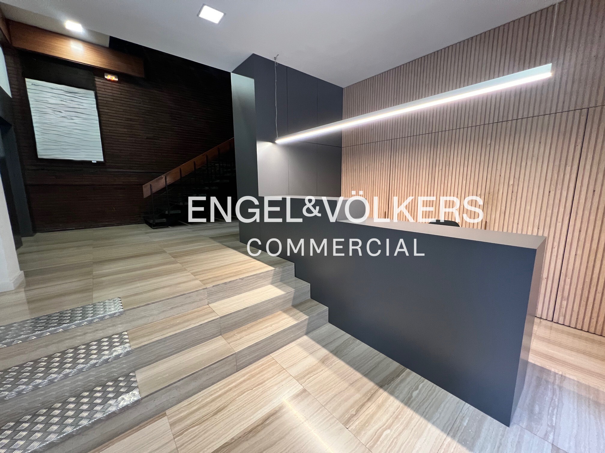 Modern office lobby with a dark gray reception desk, wood-paneled walls, tiled stairs, and a linear LED light fixture.