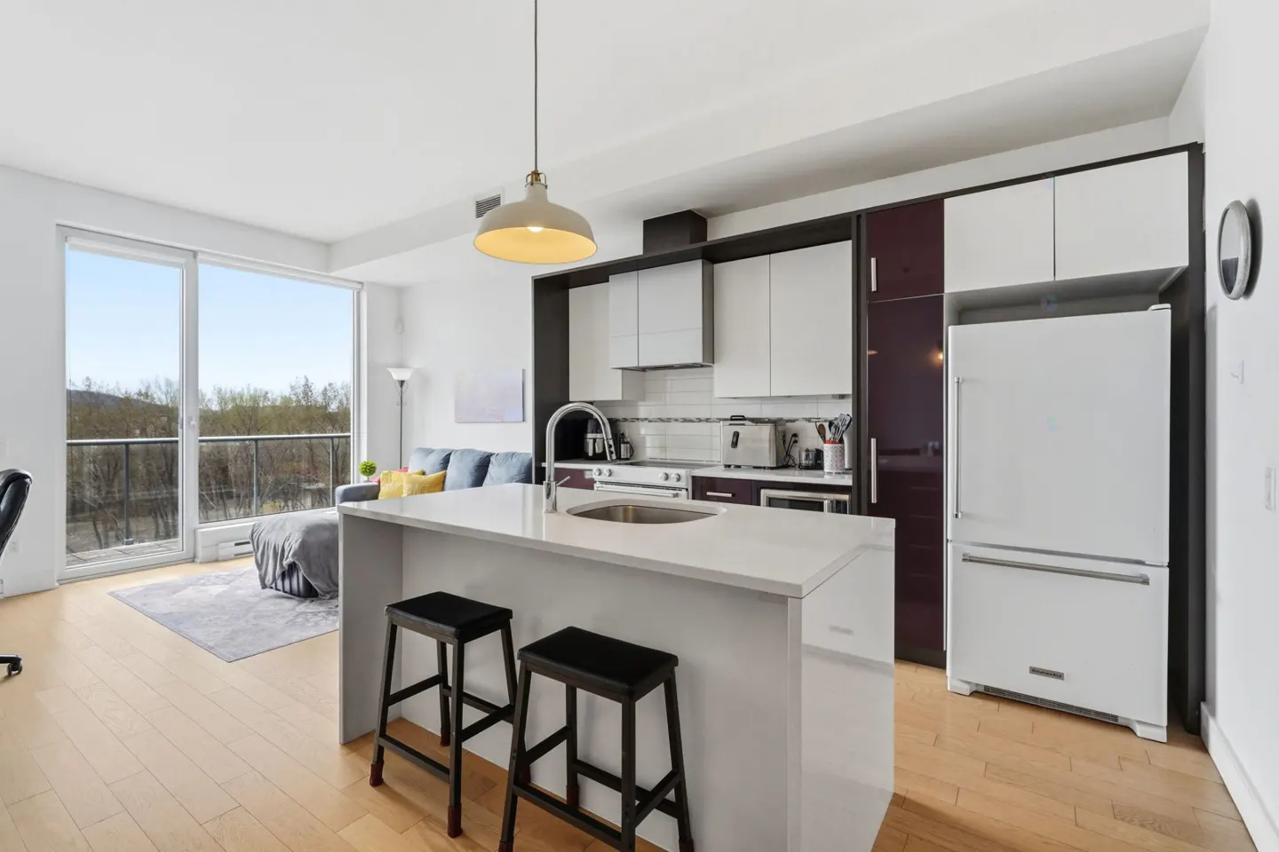 Bright, modern condo interior with a white kitchen island, black stools, and a living area with a large window.