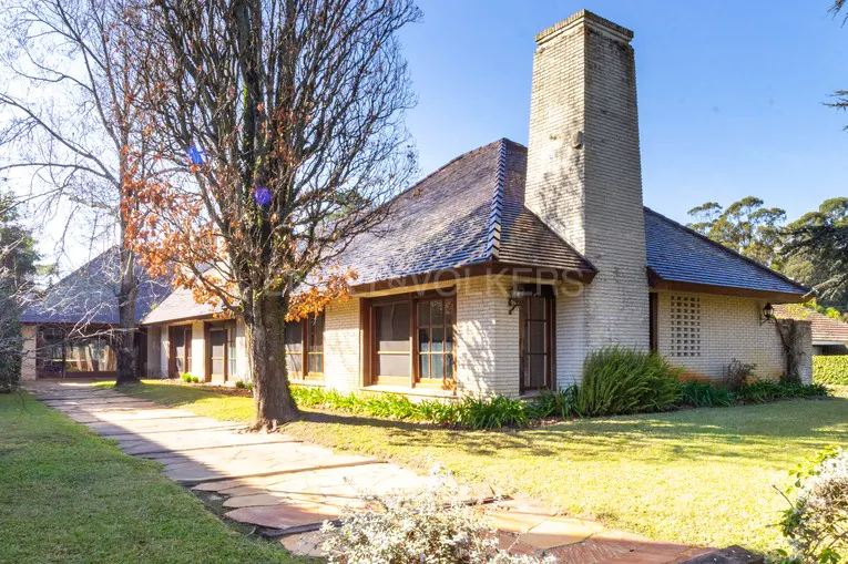 Exterior view of a one-story brick house with a tall chimney, a stone walkway, and a large tree in the front yard.