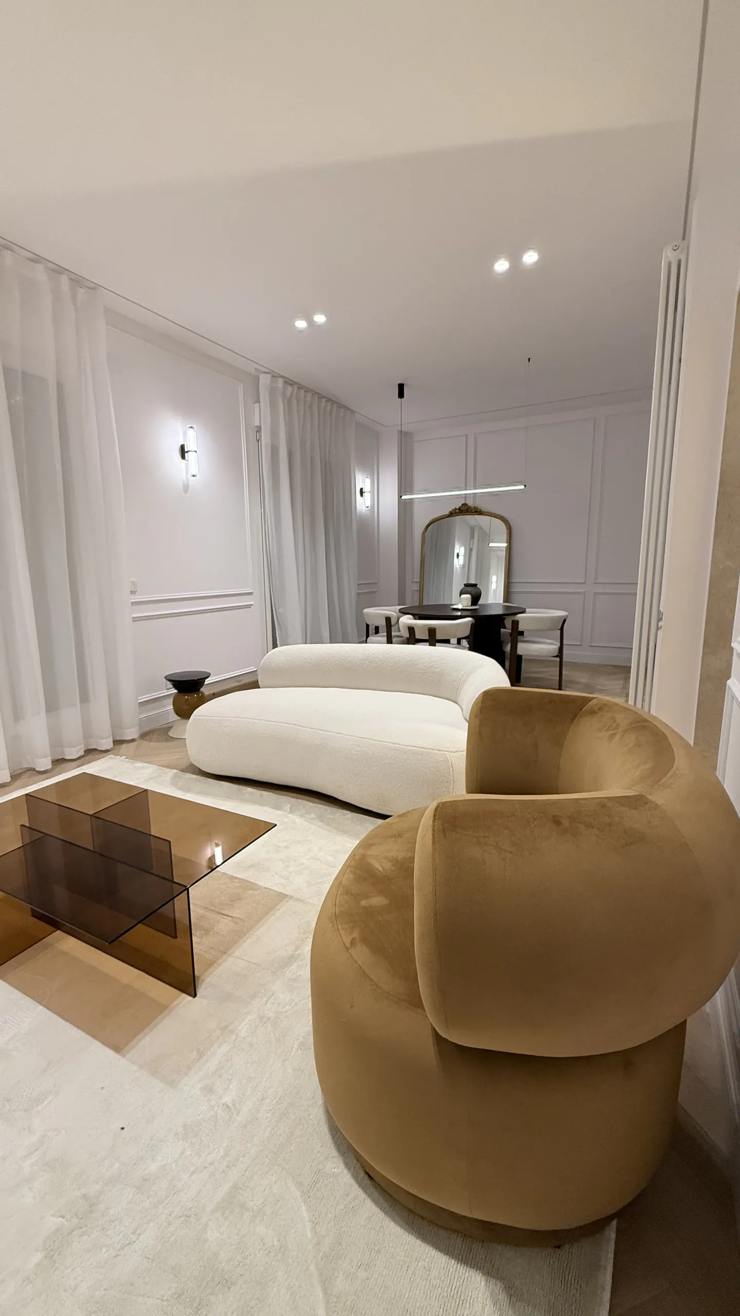 Modern living room with a curved beige sofa, a tan velvet armchair, a glass coffee table, and a dining area in the back.