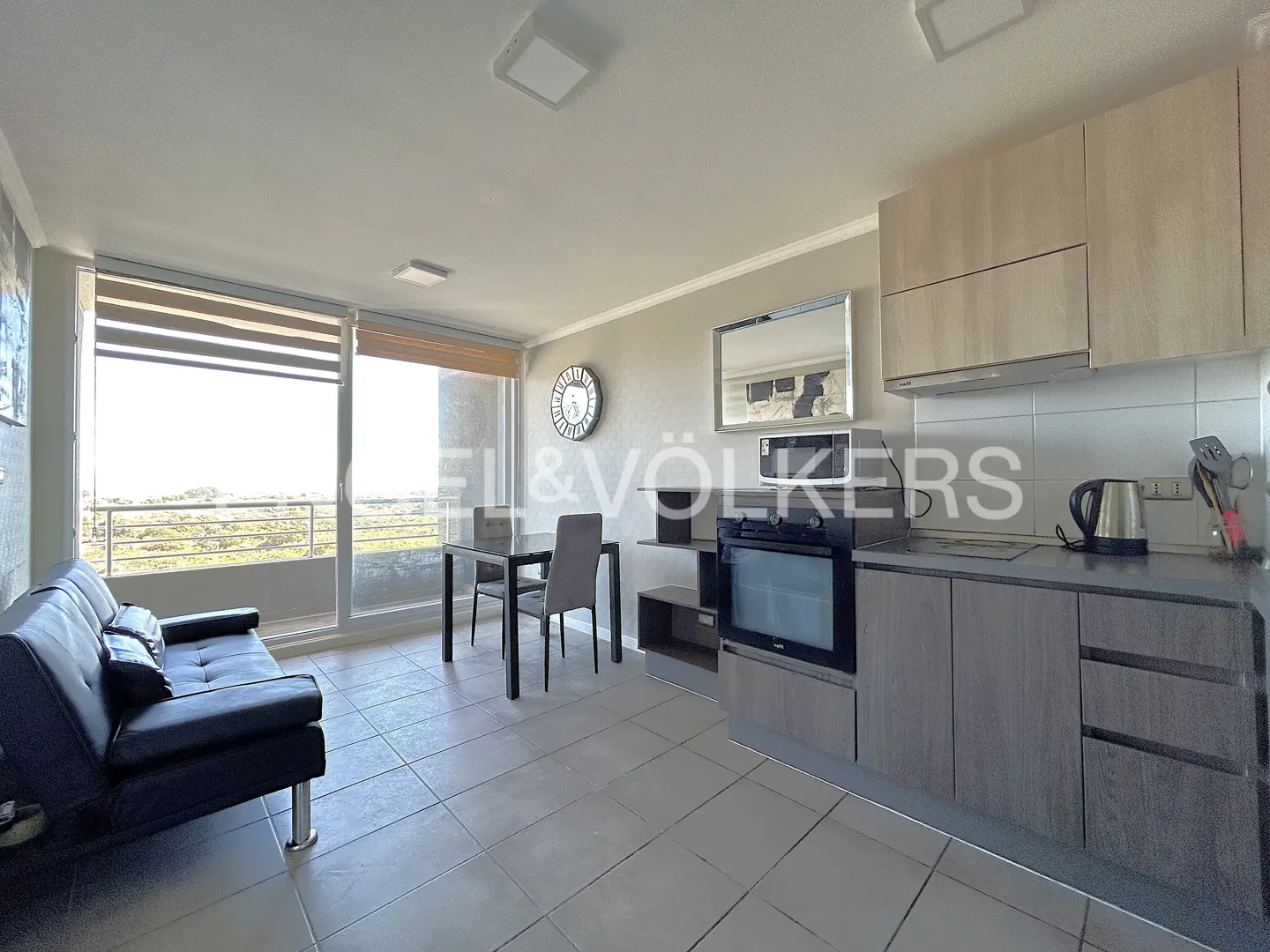 A bright apartment interior with a black sofa, dining table, and modern kitchen with gray cabinets. A large window offers a view of greenery.