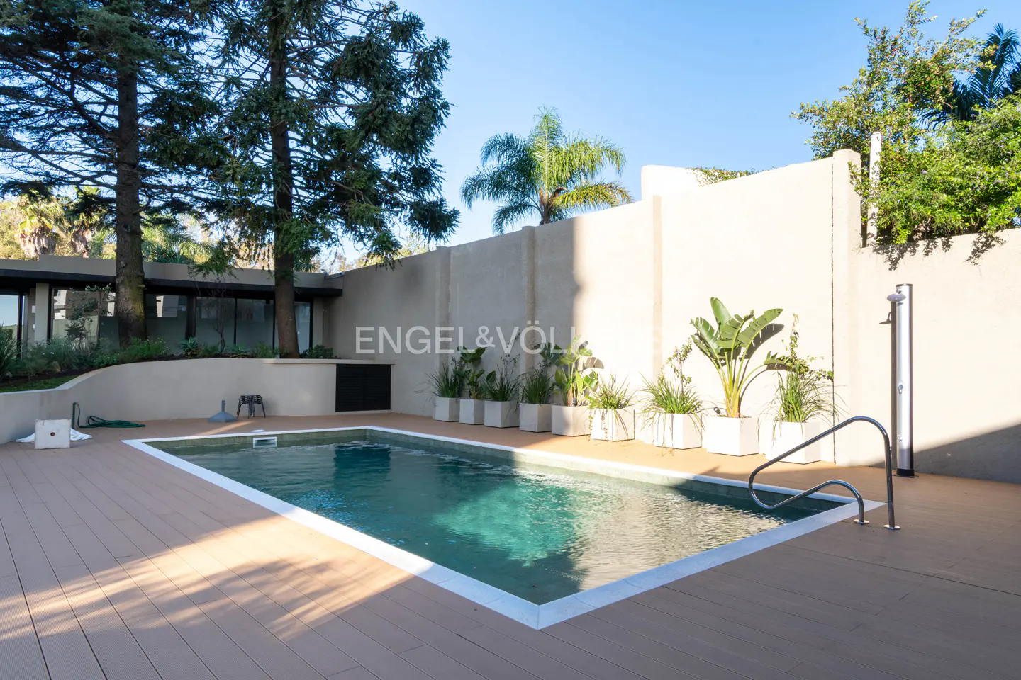 A backyard pool with a wooden deck, surrounded by a white wall and greenery. A shower stands near the pool's edge.