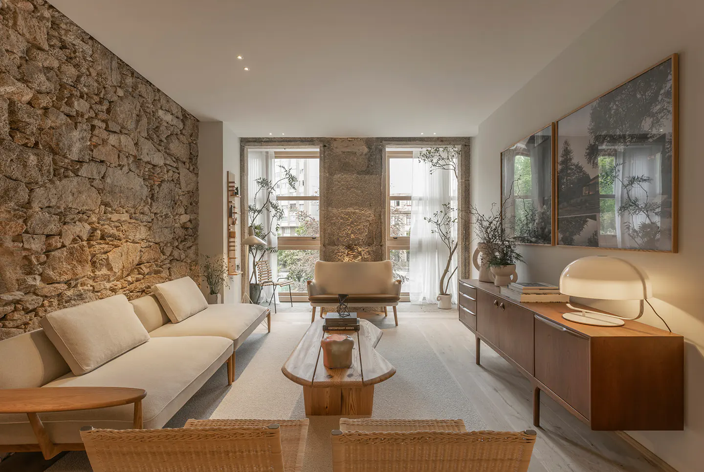 Living room with stone wall, white sofa, wood table, and cabinet. Two framed pictures hang above the cabinet. Two windows with sheer curtains.