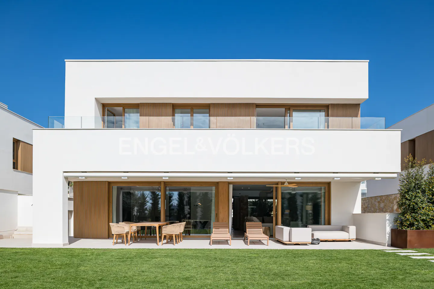 Modern white house with "Engel & Völkers" logo, patio furniture, and green lawn under a clear blue sky.