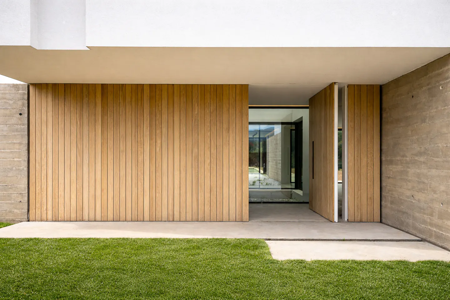 Modern home exterior with a light wood door ajar, concrete walls, and green lawn. Glass reflects the interior of the house.