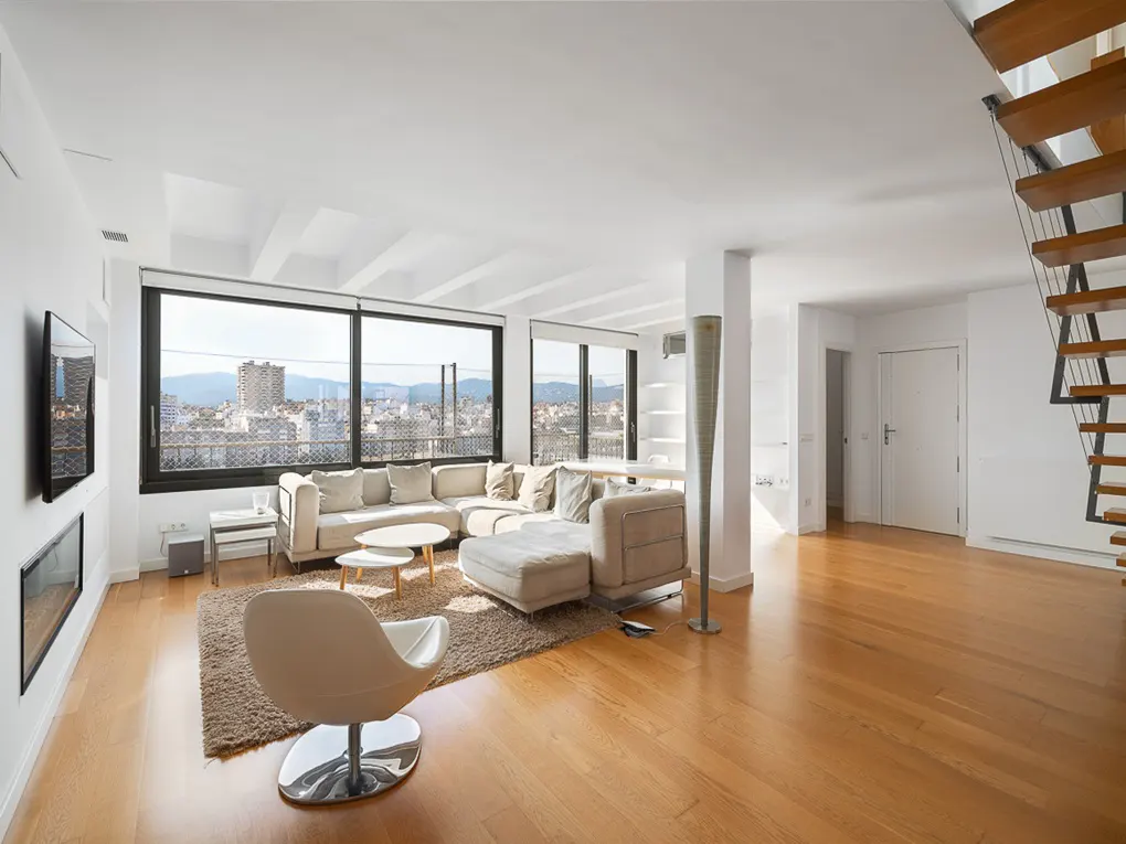 Bright, open living room with hardwood floors, a beige sectional sofa, and a large window overlooking a city. Modern staircase on the right.