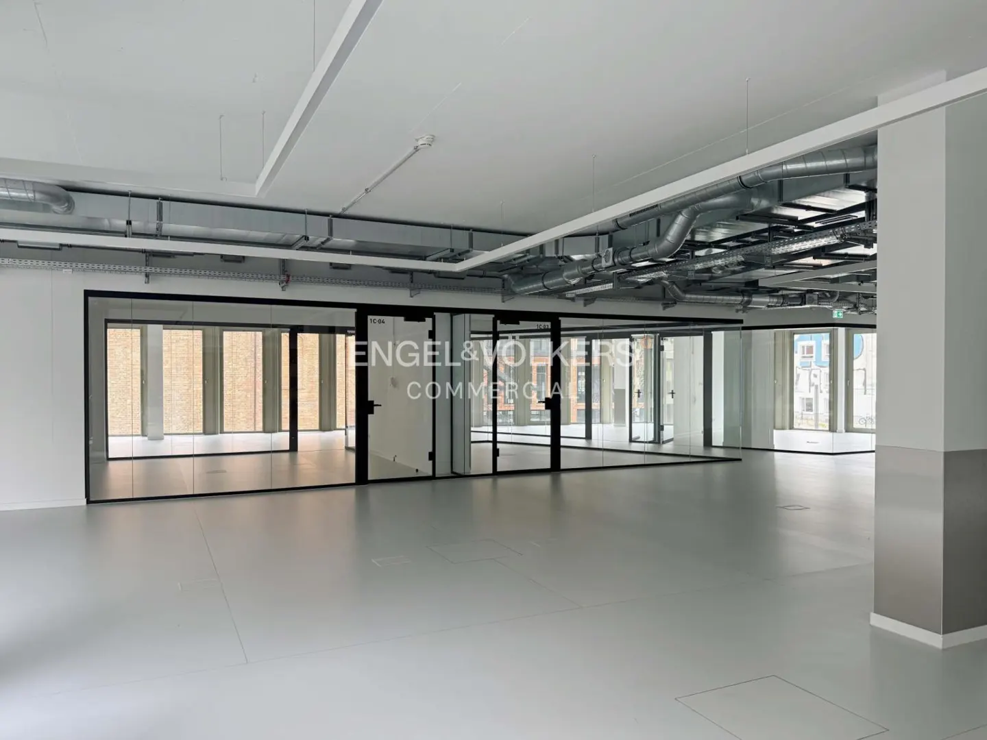 Empty office space with gray floors, white walls, and glass-walled offices. Overhead ductwork and Engel & Volkers logo on the glass.