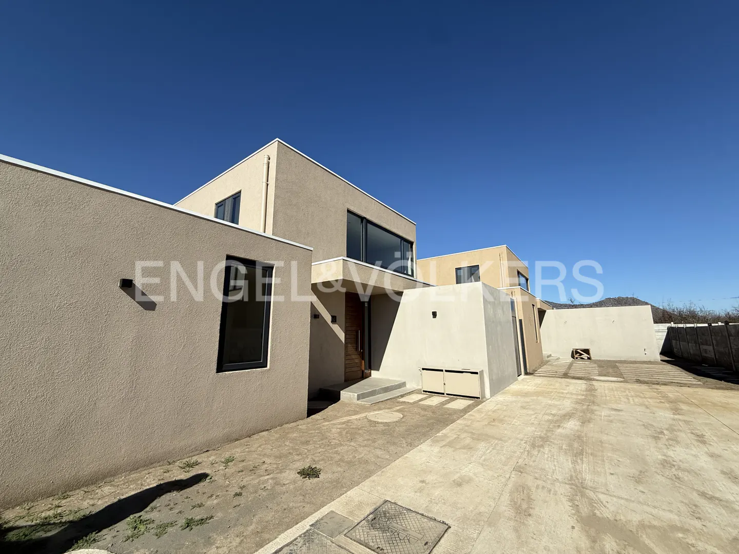 Modern beige house with black-framed windows under a clear blue sky. Concrete driveway leads to the front door.