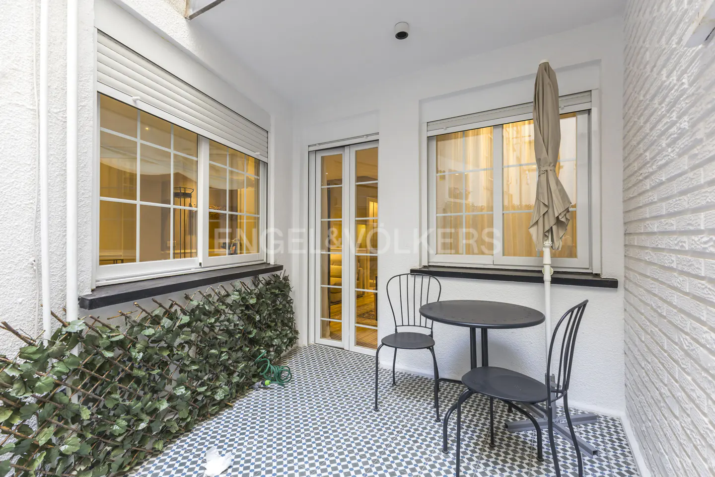 A small outdoor patio with a table, chairs, and a closed umbrella. There are windows and a door in the background. The floor is tiled.