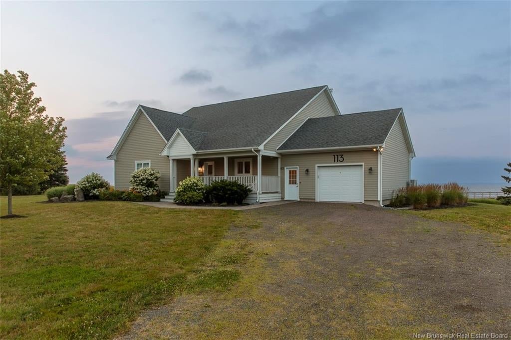 A beige house with a grey roof, front porch, and attached garage sits on a grassy lot under a cloudy evening sky. [AI-generated text]