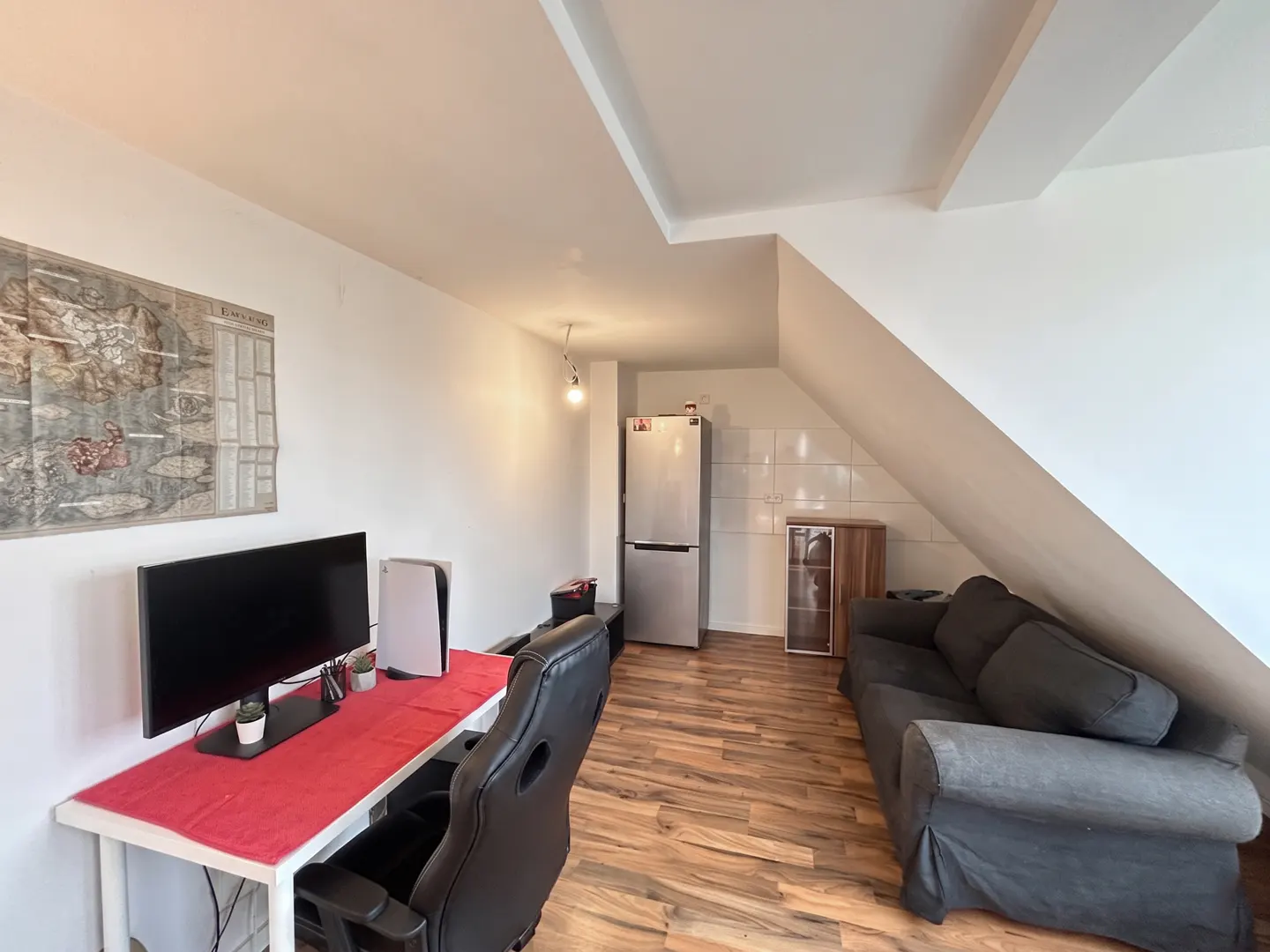 A room with a desk, monitor, PS5, and gaming chair. A gray sofa sits near a fridge and cabinet under a sloped ceiling. A world map hangs on the wall.