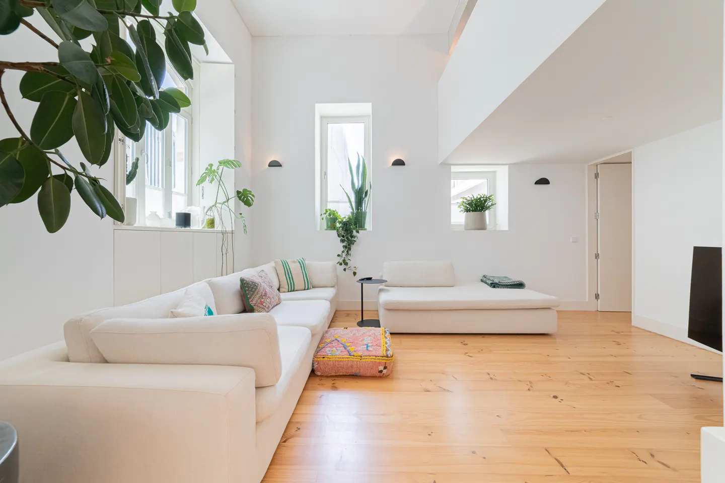Bright, modern living room with white walls, wood floors, and a large white sectional sofa. Plants are placed near the windows.